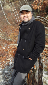 picture of author Jeffrey Nguyen in a black coat and grey baseball cap, leaning against a fallen tree. White birch trees in the background.